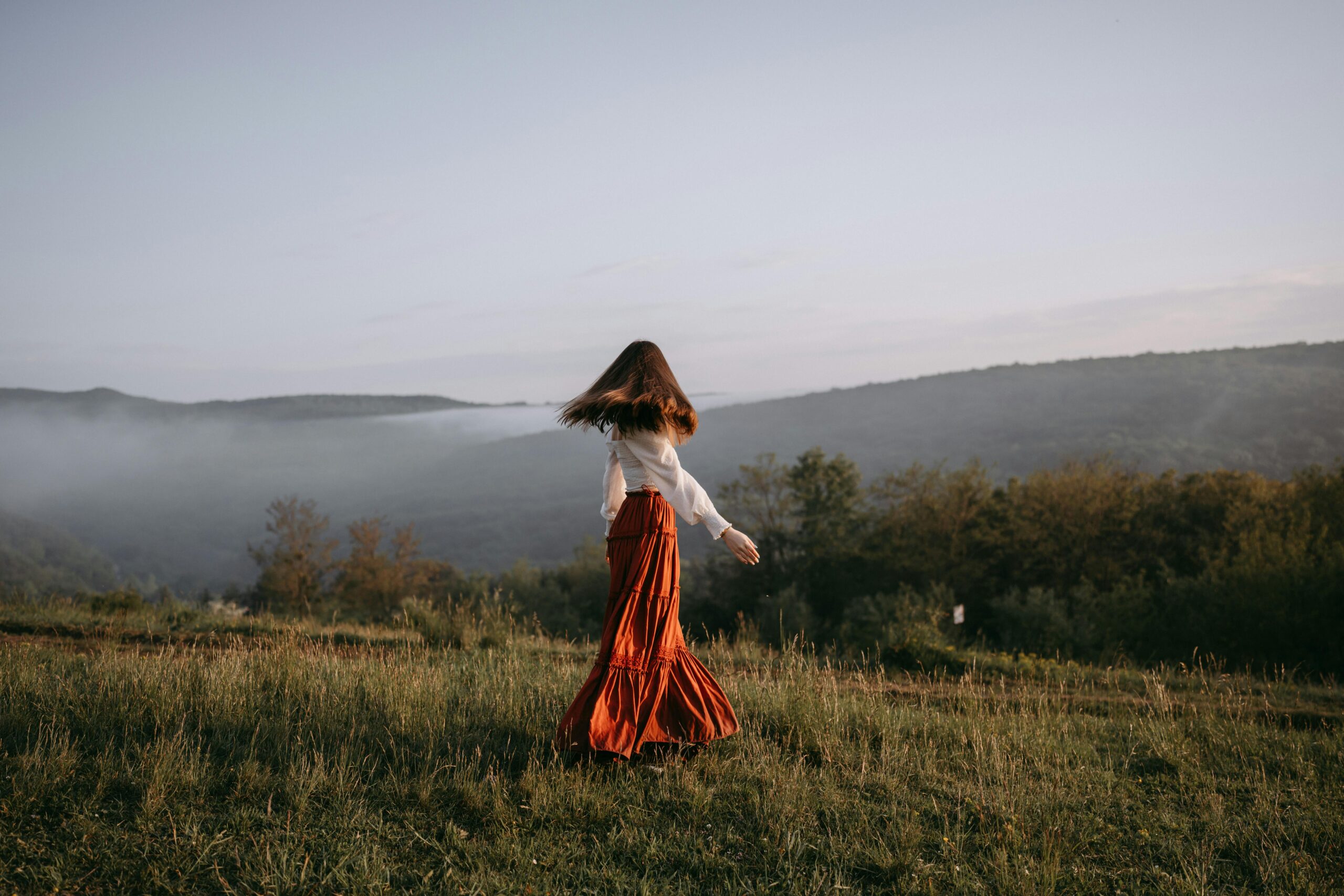 Home A woman in a flowing skirt walks through a misty meadow with hills in the background.